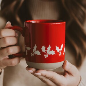 May include: A red ceramic mug with a white holly leaf design. The mug has a red handle and a beige base. The mug is held in a person's hands. The mug is suitable for hot drinks.