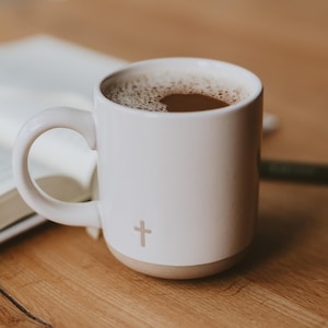 May include: White ceramic coffee mug with a cross design and a brown coffee inside. The mug is sitting on a wooden table next to a book.