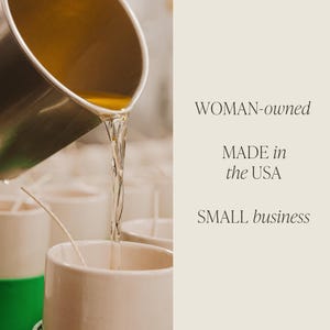 May include: A close-up of a clear liquid being poured into a white ceramic candle jar. The jar is sitting on a table with other jars. The text "WOMAN-owned MADE in the USA SMALL business" is visible in the background.