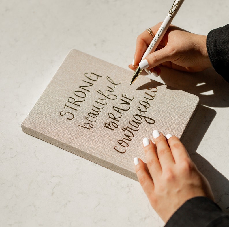 May include: A beige linen journal with the words "STRONG, beautiful, BRAVE, courageous" written in gold calligraphy. A hand is holding a white pen with the words "LOVE YOURSELF" written on it.
