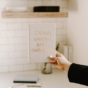 May include: A beige linen journal with gold lettering that reads "STRONG beautiful brave courageous".