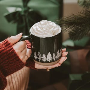 May include: A black ceramic mug with a white design of trees around the rim. The mug is filled with a creamy beverage topped with whipped cream.