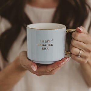 May include: A light blue ceramic mug with gold accents on the handle and rim. The mug has the text "IN MY ENGAGED ERA" in gold lettering, with a small ring graphic. The mug is held by a person.