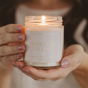 Może przedstawiać: Zapalone świeca w szklanym słoiku z etykietą z napisem "MANGO and COCONUT". Etykieta zawiera r&oacute;wnież tekst "SWEET WATER DECOR". Świeca jest trzymana w dłoniach osoby.