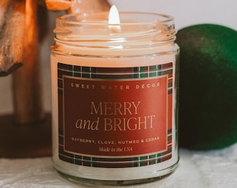Könnte beinhalten: Eine brennende Glas-Kerze mit einem roten Etikett mit der Aufschrift "MERRY and BRIGHT". Das Etikett hat einen karierten Rand und listet die Düfte auf: Lorbeerbeere, Nelke, Muskatnuss und Zeder. Die Kerze wird in den USA hergestellt.