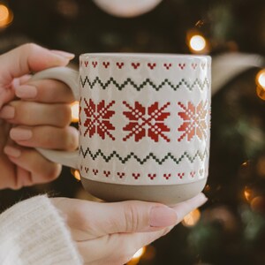 May include: A ceramic mug with a white base and a textured surface featuring a repeating red and green Nordic-style snowflake and zigzag pattern. The bottom section of the mug is a solid matte gray, and it includes a curved handle on the side.