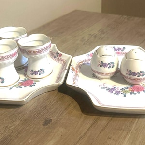 May include: A set of two serving trays with matching egg cups and salt and pepper shakers. The white porcelain pieces feature a floral and leaf design with pink, purple, and gold accents. The egg cups are on a tray with a scalloped edge.