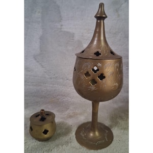 May include: A tall brass incense burner with a pointed lid and a matching small round brass container. Both items feature cut-out clover-shaped patterns and delicate etched line details across their surfaces, standing on circular bases.