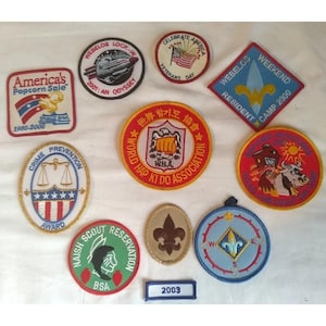 May include: A collection of embroidered patches with various designs and text. Patches include "America's Popcorn Sale," "Webelos Lock-In," "Celebrate America Veterans Day," and "Crime Prevention Award." Other patches feature a compass, a fleur-de-lis, and a Native American profile.