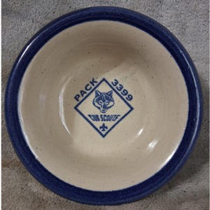 May include: A round ceramic bowl with a speckled cream interior and a dark blue rim. The center features a blue diamond-shaped logo with a wolf head, the text PACK 3399, the words CUB SCOUTS, and a fleur-de-lis symbol printed on the bottom surface.