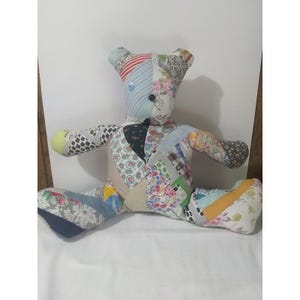 Könnte beinhalten: Ein handgefertigter Patchwork-Teddybär mit verschiedenen Stoffmustern und -farben. Der Bär hat Knopfaugen und eine Knopf-Nase. Die Arme und Beine sind ausgestreckt, und er sitzt auf einer weißen Oberfläche.