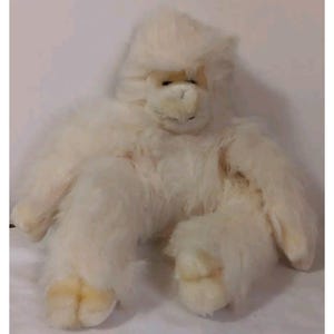 May include: A plush, off-white gorilla toy with a soft, furry texture. The toy has a detailed face with a light brown snout and small, dark eyes. Its arms and legs are outstretched, and the feet are a lighter shade of cream.