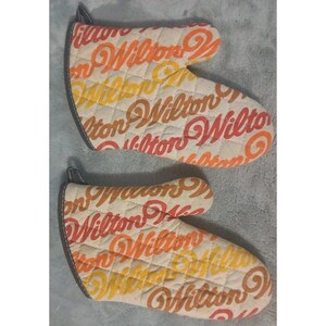 May include: Two quilted oven mitts featuring the word "Wilton" in orange, yellow, and red script. They have a diamond pattern and a dark grey trim. These mitts are designed for use in the kitchen.