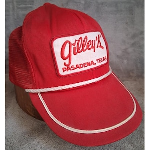 May include: A bright red trucker hat featuring a white rectangular patch on the front with the red embroidered text Gilley's PASADENA, TEXAS. The cap includes a white braided cord across the base of the crown and a matching white trim on the brim.