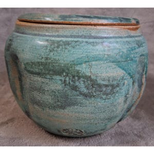 Op de afbeelding: Een ronde keramische pot met deksel, voorzien van een gespikkeld blauw-groen glazuur met zichtbare horizontale penseelstreken. De pot heeft een brede, licht taps toelopende vorm met subtiele verticale inkepingen langs de zijkanten.