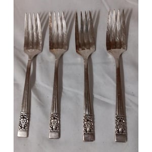 May include: Four silver forks with tines and decorative handles. The handles feature a detailed floral design at the base. The forks are arranged on a white surface.