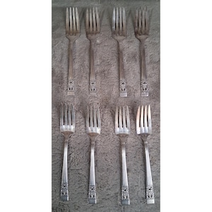 Pode incluir: Um conjunto de oito garfos de metal prateado dispostos em duas filas de quatro. Cada garfo apresenta quatro dentes e um cabo com um padrão de ranhuras verticais e um motivo floral decorativo em relevo na base do cabo.