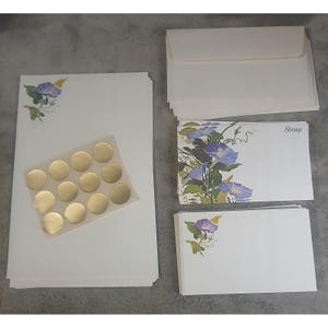 May include: Stationery set featuring white paper, envelopes, and notecards. The paper and cards are adorned with a floral design of purple and blue morning glories. Includes a sheet of gold circle stickers. The word "Stamp" is printed on one envelope.