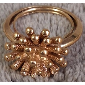May include: A gold-toned ring featuring a central cluster of numerous radiating, textured stems topped with small, smooth, spherical beads, creating a dense, starburst-like focal point on a simple, polished circular band.