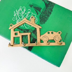 May include: A gold-toned metal key holder shaped like a house with a car attached. The house has a chimney and four hooks. The car has four wheels and a rounded shape.