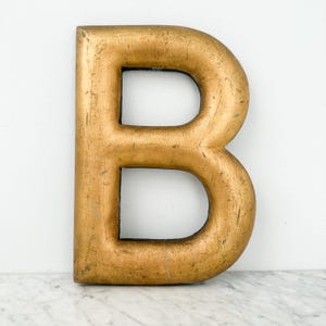 May include: A large, three-dimensional capital letter B with a distressed gold-leaf finish. The surface shows visible signs of wear, including fine scratches and subtle texture, with a dark interior edge visible within the letter's two openings.