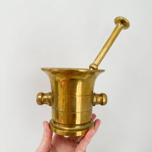 May include: A vintage brass mortar and pestle set. The mortar has a flared rim, two side handles, and a round base. The pestle is long and cylindrical with a rounded end. The set is a shiny gold color.