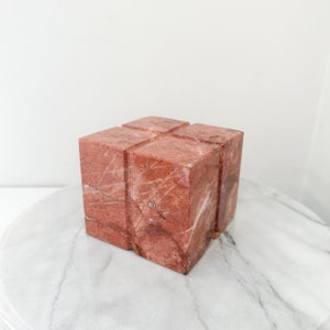 May include: A solid red marble block sculpture featuring a cross-shaped groove carved into the top surface. The stone displays natural white veining throughout its polished, cubic structure, showcasing the unique texture and color variations of the material.