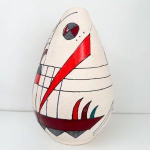 May include: Cream-colored, teardrop-shaped ceramic vase with abstract geometric designs. Features a large red diagonal stripe, black lines, and red, gray, and teal accents. The vase has a textured surface and a unique, artistic aesthetic.