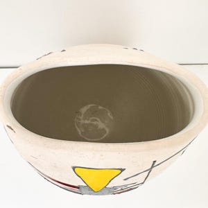 May include: A cream-colored ceramic bowl with a wide opening. The bowl features a yellow triangle and gray and black geometric designs. The interior is a light brown color. The bowl is likely used for decorative purposes.