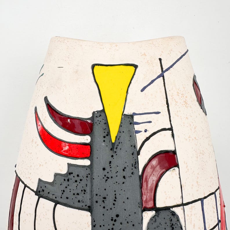 May include: A cream-colored ceramic vase with a geometric design. The vase features a yellow triangle, red curved shapes, and black lines. The design also includes gray and black speckled areas.