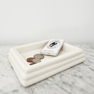 May include: An off-white ceramic tray with a tiered design, holding coins and a small white box. The box is labelled "Forest Creek Golf Club Pinehurst NC". The tray sits on a marble surface. The tray's dimensions are not visible.