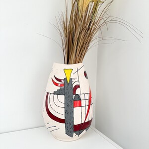 May include: Cream-colored ceramic vase with abstract geometric designs in red, gray, and black. A yellow triangle and curved lines accent the vase. Tall, dried grasses are arranged inside the vase, creating a decorative display.