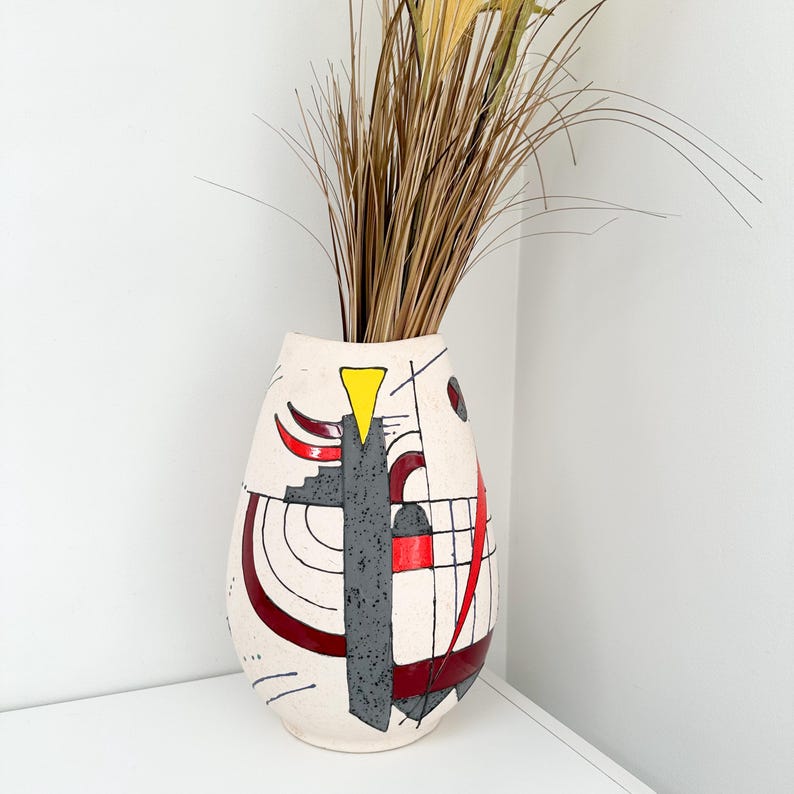 May include: Cream-colored ceramic vase with abstract geometric designs in red, gray, and black. A yellow triangle and curved lines accent the vase. Tall, dried grasses are arranged inside the vase, creating a decorative display.