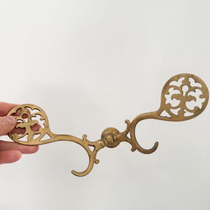 May include: A double-sided brass wall hook featuring two symmetrical arms that extend outward. Each arm terminates in a flat, circular plate with an intricate, openwork cutout pattern. The central mounting point is a small, circular brass base.