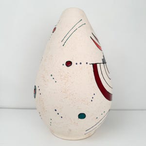 May include: A cream-colored, teardrop-shaped ceramic vase with abstract painted details. The vase features red, teal, and black accents, including dots and curved lines. The surface has a textured appearance.