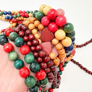 Puede incluir: Una colección de collares y pulseras de cuentas de madera de colores. Las cuentas son de varios colores, incluyendo rojo, verde, amarillo y marrón. Algunas pulseras presentan cuentas de madera en forma de corazón. La joyería se muestra sobre un fondo blanco.