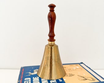 May include: A brass bell with a wooden handle. The bell is sitting on top of a blue book with the text "A FIELD GUIDE TO ANIMAL TRACKS BY THE PETERSON FIELD GUIDE SERIES".