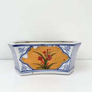 May include: A rectangular ceramic planter with a white and blue geometric pattern. The center features a gold-colored design with red flowers and green leaves. The planter has a curved base.