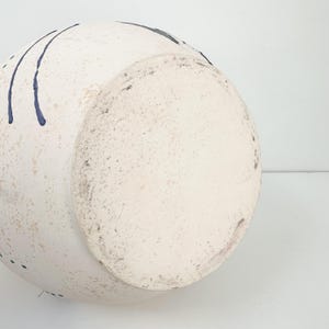 May include: A white ceramic vase with a rounded shape and a textured surface. The vase features dark blue drip-like accents and a speckled appearance. The bottom of the vase is visible, showing a lighter, unglazed area.