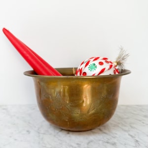 Puede incluir: Un cuenco de latón vintage con diseños florales grabados contiene dos velas rojas y un adorno navideño. El adorno es blanco con detalles rojos y verdes. El cuenco está sobre una superficie de mármol.