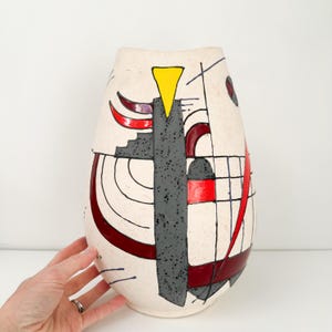 May include: A cream-colored ceramic vase with an abstract design. The vase features a central gray vertical element, red curved shapes, and black lines. A yellow triangle and red square add pops of color to the design. The vase has a textured surface.