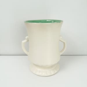 May include: Cream-colored ceramic vase with a green interior rim and two curved handles. The vase has a pedestal base with a scalloped edge. A decorative piece for home decor.