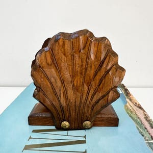 May include: A wooden napkin holder shaped like a seashell with a brown wood finish. The holder has two brass accents on the bottom.