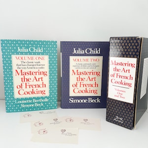 May include: Three cookbooks by Julia Child, titled "Mastering the Art of French Cooking." Volume One is turquoise, Volume Two is navy, and a boxed set of Volumes One and Two. Several small cards with a heart and floral design are in front.