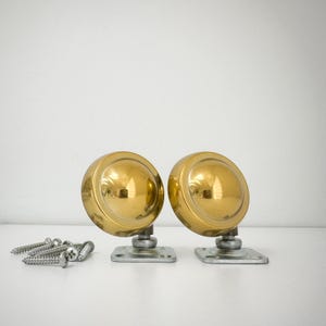 May include: Two vintage brass casters with a shiny, reflective finish. Each caster has a rounded, dome-shaped top and a square base. A small pile of silver screws is visible to the left. These casters are likely for furniture.