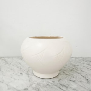 Puede incluir: Un jarrón de cerámica redondo de color crema con una abertura ancha y una base corta y circular. El jarrón presenta un sutil patrón de hojas repetido alrededor de su cuerpo. El interior del jarrón es de color marrón claro. El jarrón está sobre una superficie de mármol.