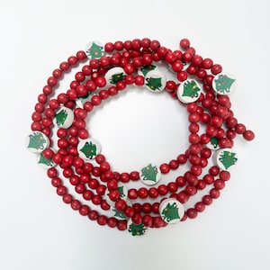 May include: A festive Christmas garland featuring red wooden beads and white circular accents. Each accent displays a green Christmas tree design. The garland is arranged in a circular pattern against a white background.