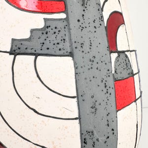 May include: A close-up of a ceramic vase with a geometric design. The vase features a white background with black outlines, gray shapes, and red accents. The surface has a speckled texture.