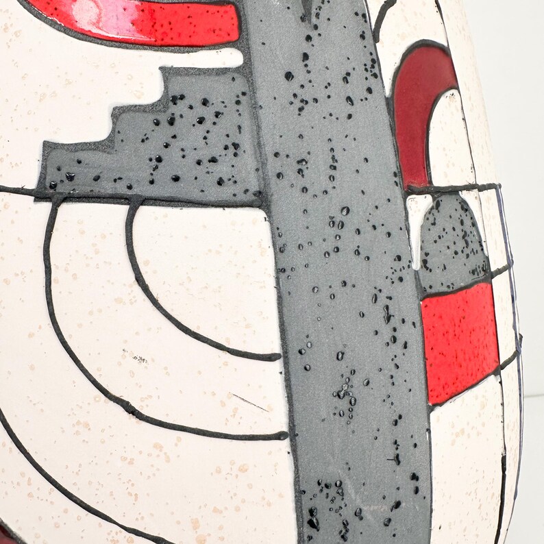 May include: A close-up of a ceramic vase with a geometric design. The vase features a white background with black outlines, gray shapes, and red accents. The surface has a speckled texture.