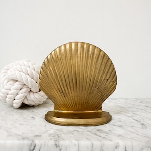 May include: A gold-toned metal seashell-shaped object with a fan-like ribbed texture and a smooth, curved base. The shell features vertical ridges radiating from the bottom center to the rounded top edge, creating a distinct scalloped silhouette.